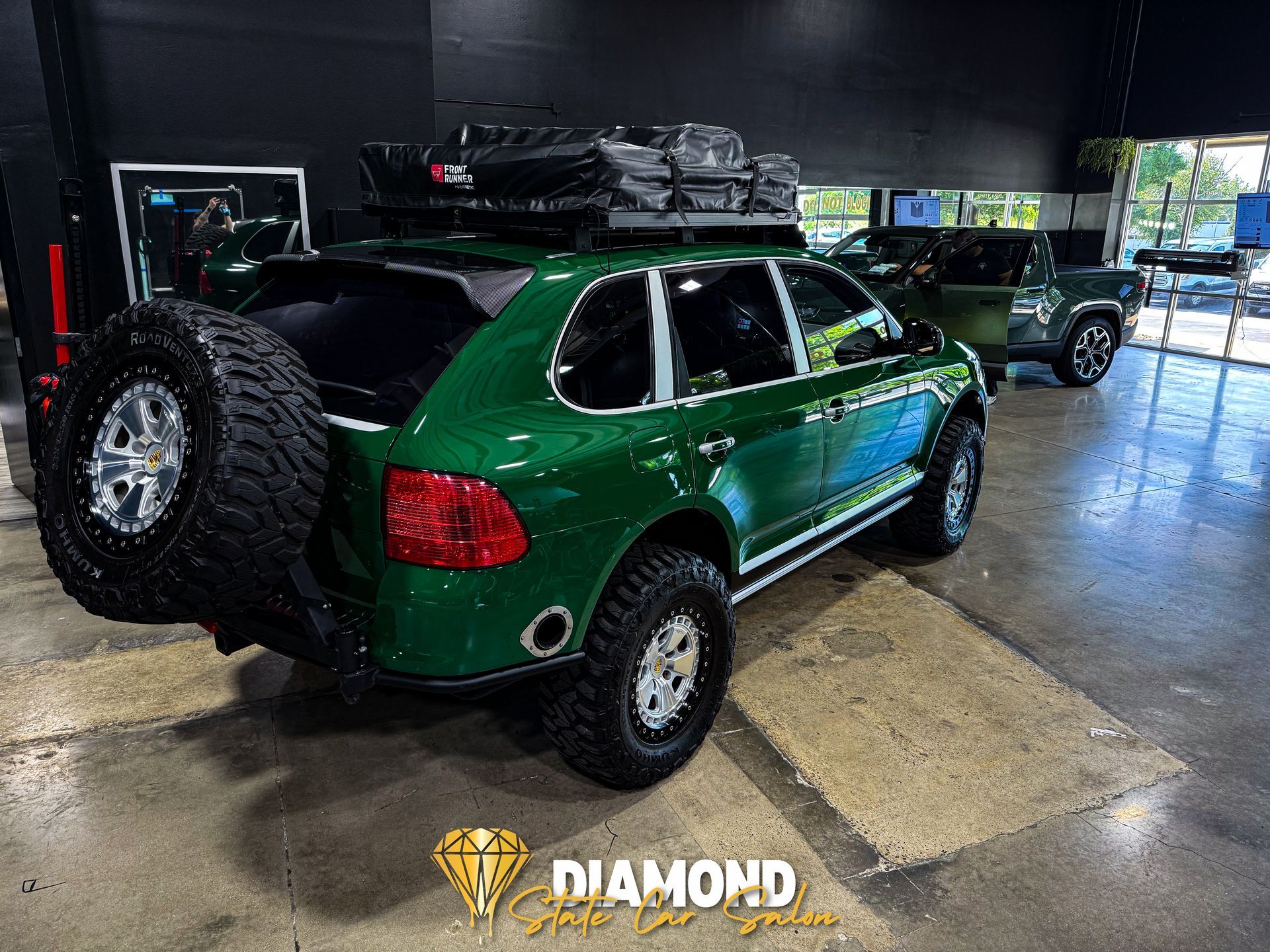 Green lifted Porsche Cayenne SUV with a spare tire, roof rack, and off-road tires, in a showroom.