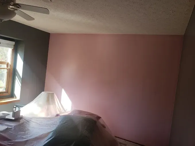 A room with a pink wall, a gray wall, and a window. A lamp is covered with plastic.