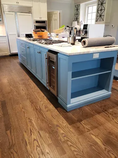 Large blue kitchen island with a wine fridge, shelves, and white countertop in a kitchen with hardwood floors.
