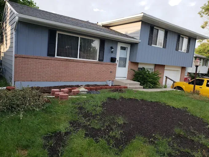A two-story blue house with red brick and a yellow truck in front of it. Dirt and bricks are in the yard.