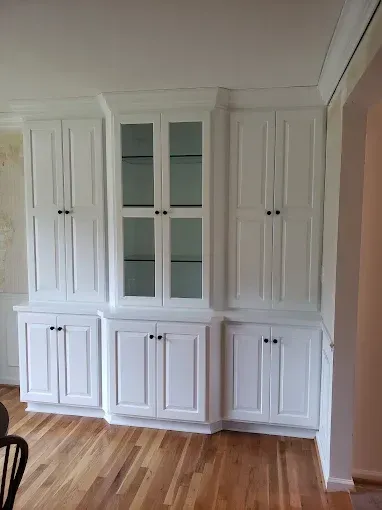 White built-in cabinets with glass-door center, against a wall in a room with hardwood floors.