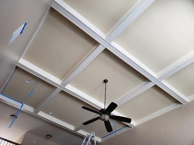 Beige ceiling with white beams forming a grid. A black ceiling fan hangs in the center, and blue wires are visible.
