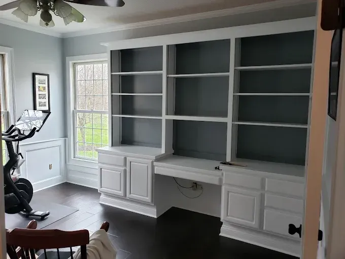 Built-in white bookshelves with a desk in a room. A Peloton bike sits near a window. Dark wood floor.