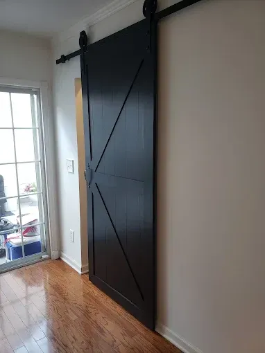 Black barn door on a sliding track, wood floor, white wall, glass window with outdoor view.