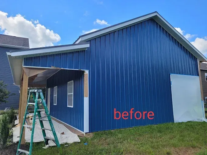 Blue metal-sided building with white trim, unfinished wood, and a green ladder on a grassy hill; "before" text.