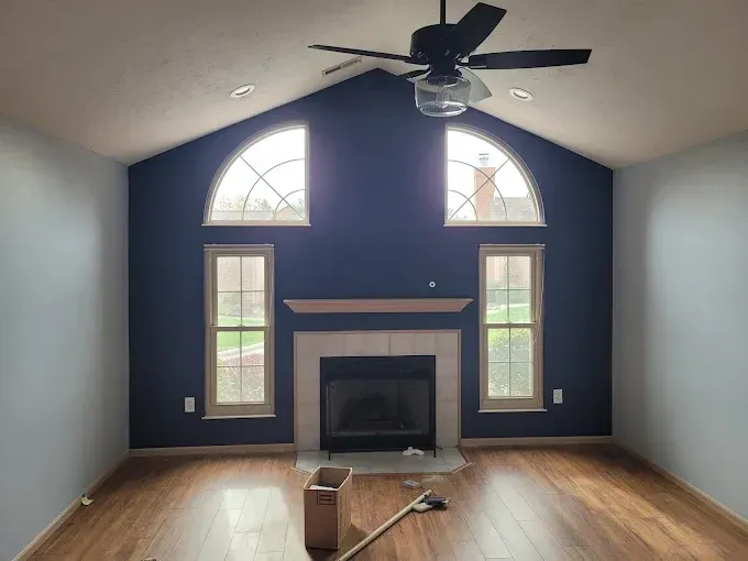 Living room with a dark blue accent wall, fireplace, arched windows, ceiling fan, and hardwood floor.