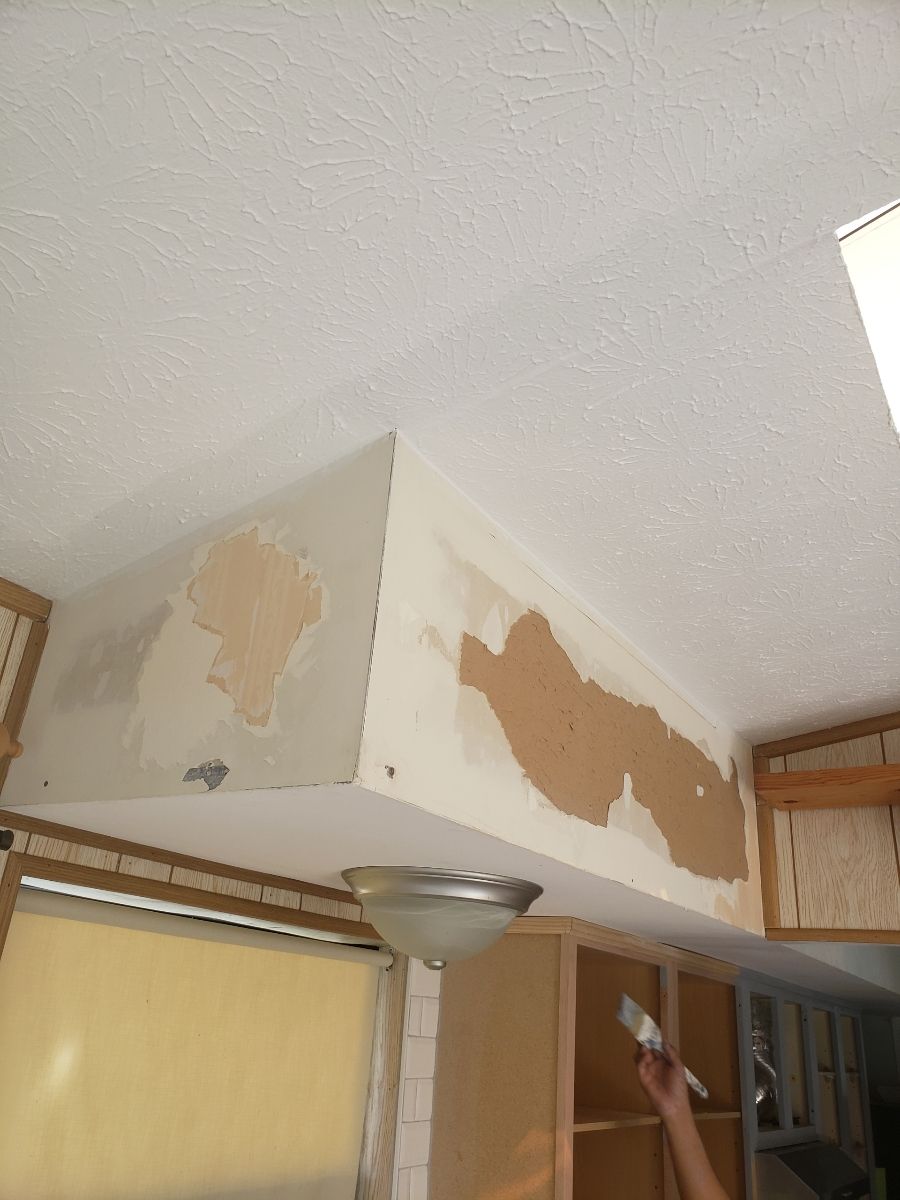 Ceiling repair above kitchen cabinets, patching drywall with visible brown patches and person painting.
