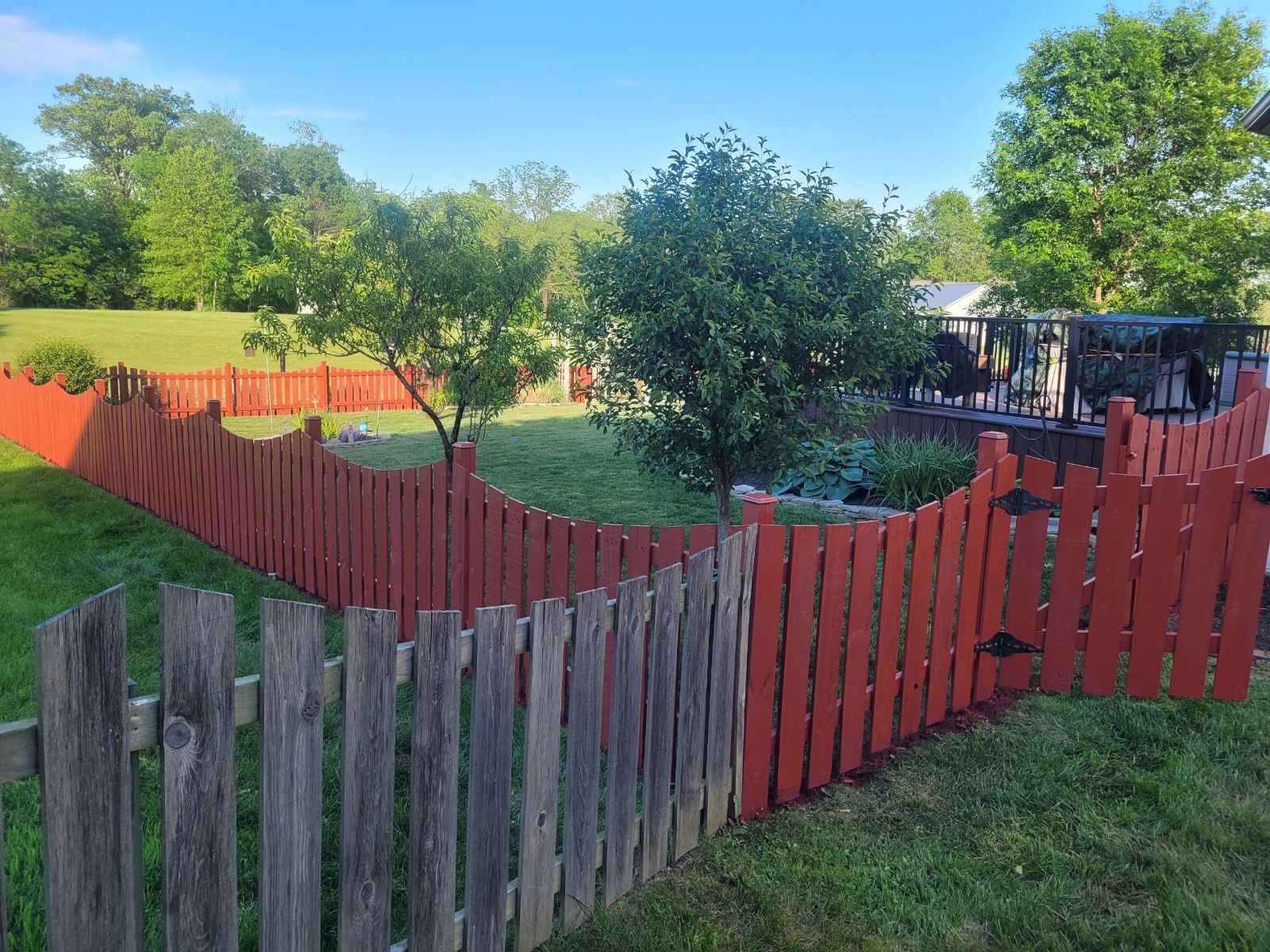 Red and weathered wooden fence encloses a green yard with trees and a building in the background.