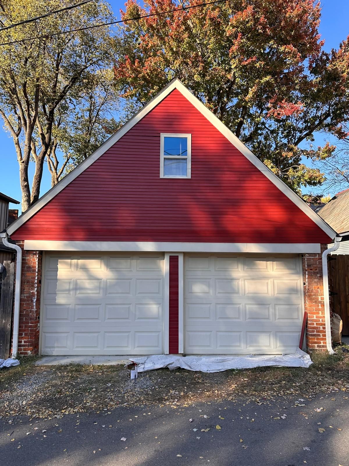 Red-gabled garage with two white doors, a small window, and brick accents. Fall foliage in the background.