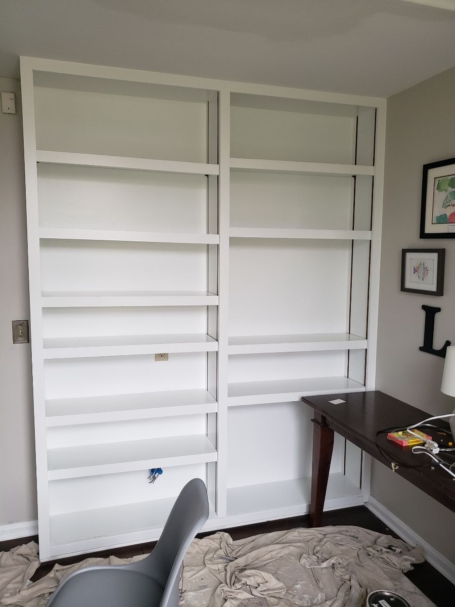 White built-in bookshelves against a white wall; desk on right side, chair and paint supplies in the foreground.