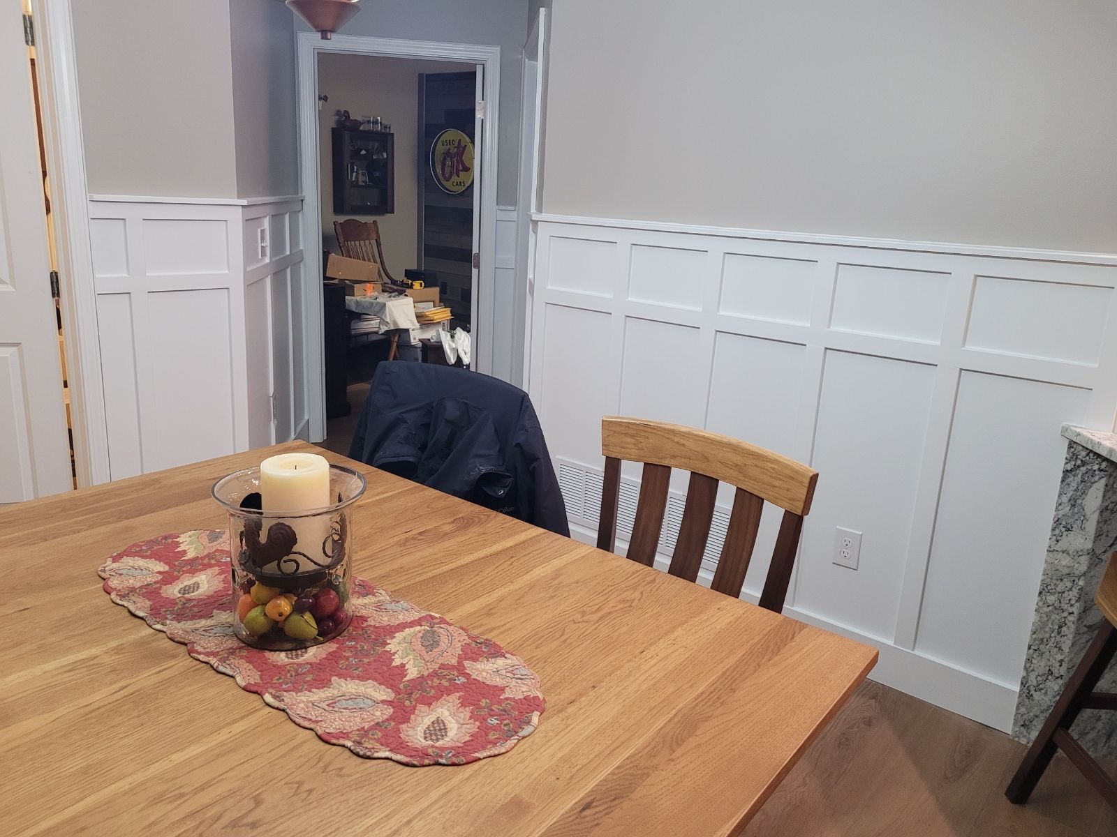 Wooden table with candle and runner, chair in dining room with white wainscoting and doorway in background.