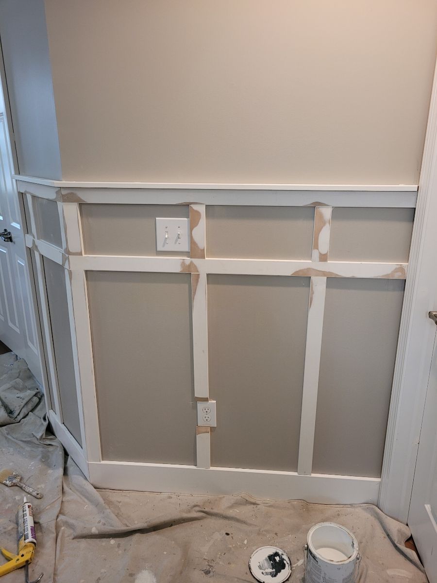 Wall with wainscoting under construction; white trim on tan wall; electrical outlets visible; paint supplies on floor.