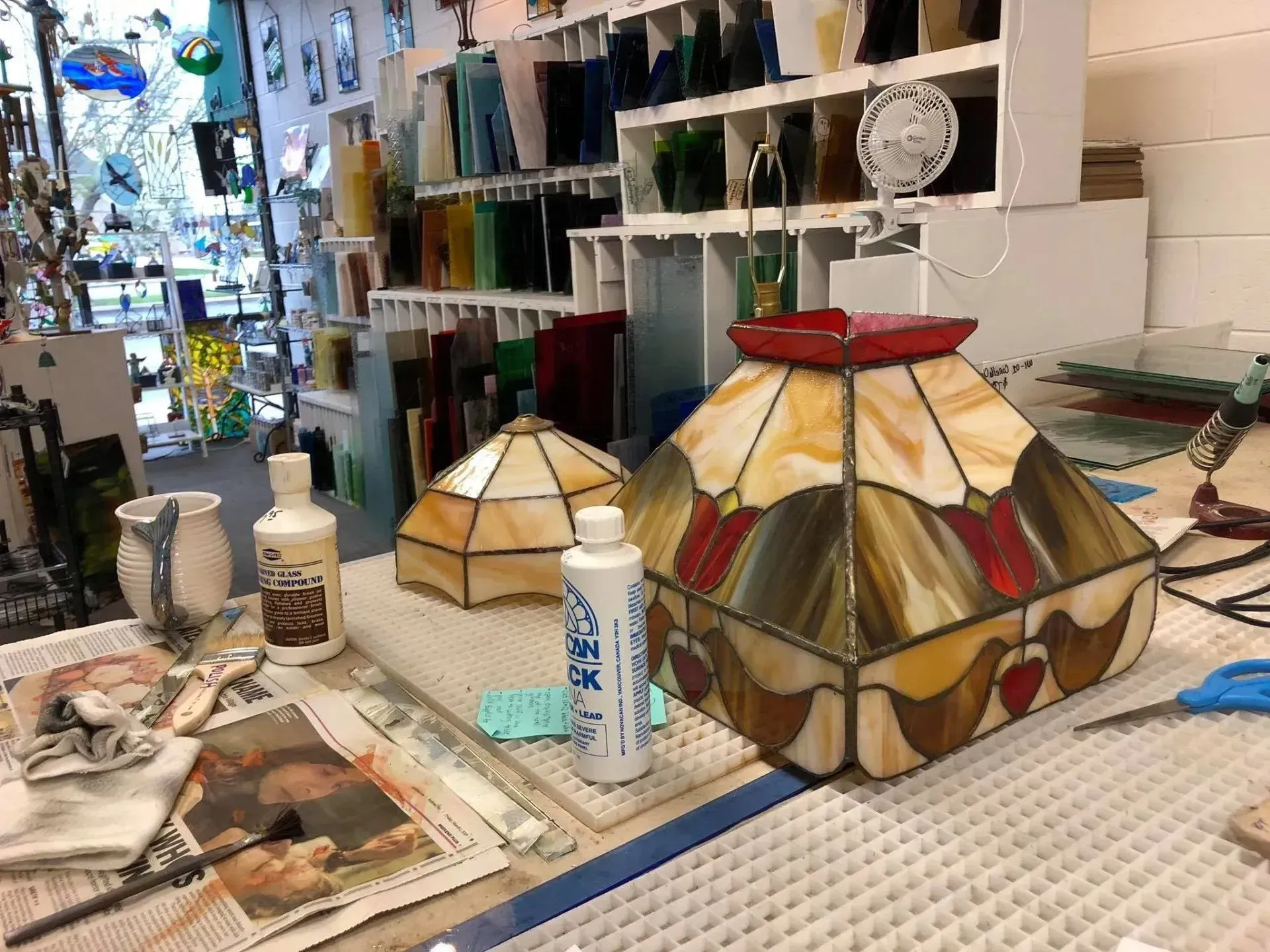 A craft studio table with stained glass lampshades, glass shelves, and various crafting tools.