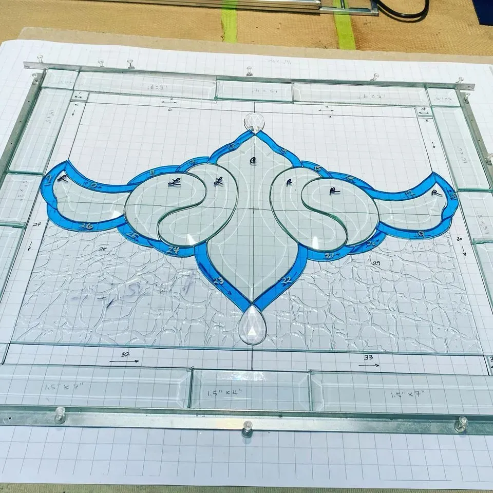 Stained glass panel in progress, light blue and clear design. Birds with blue outlines, set on grid paper, surrounded by metal framing.