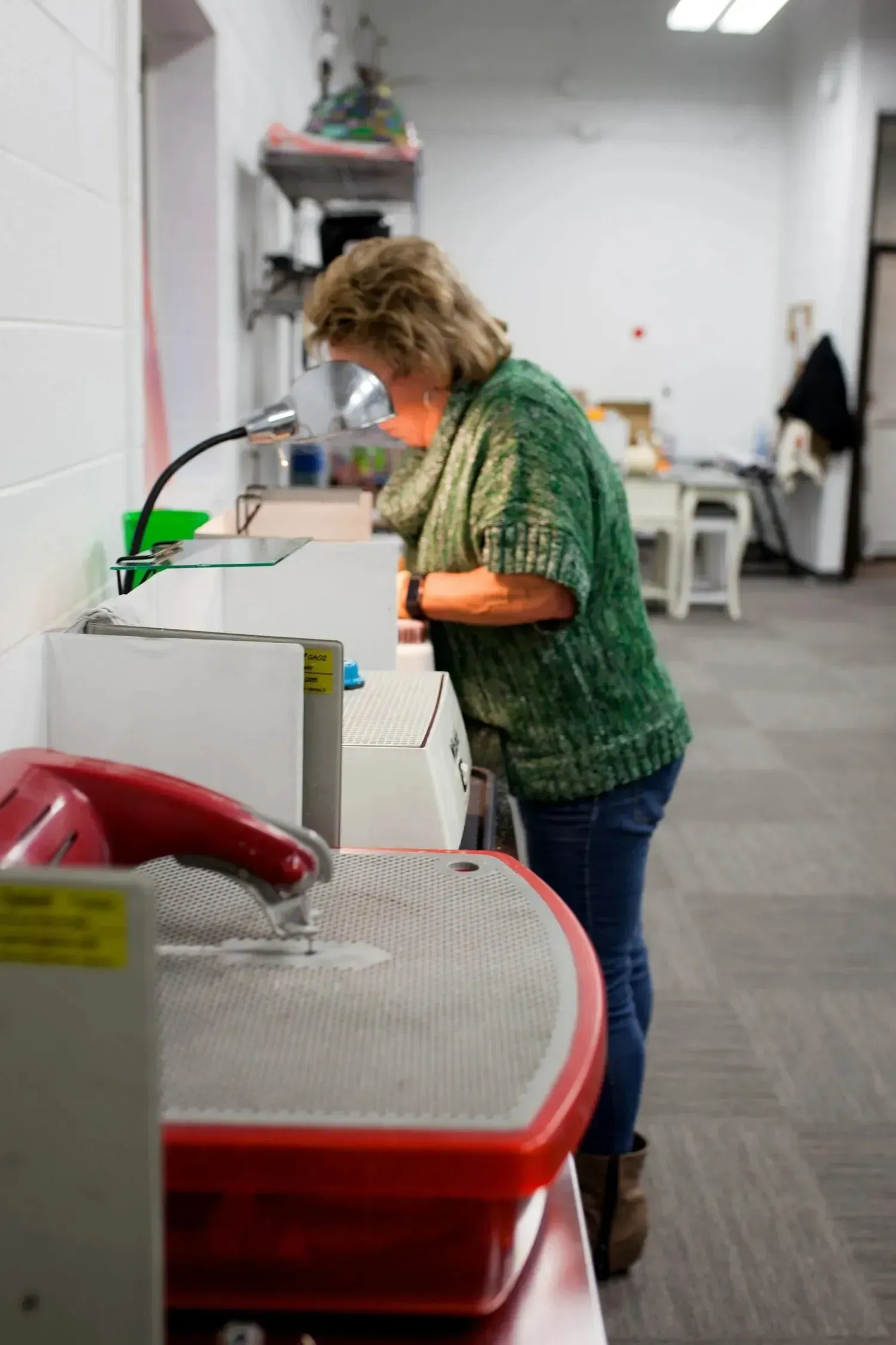 Woman working at a lapidary machine in a workshop, wearing a green sweater and jeans.