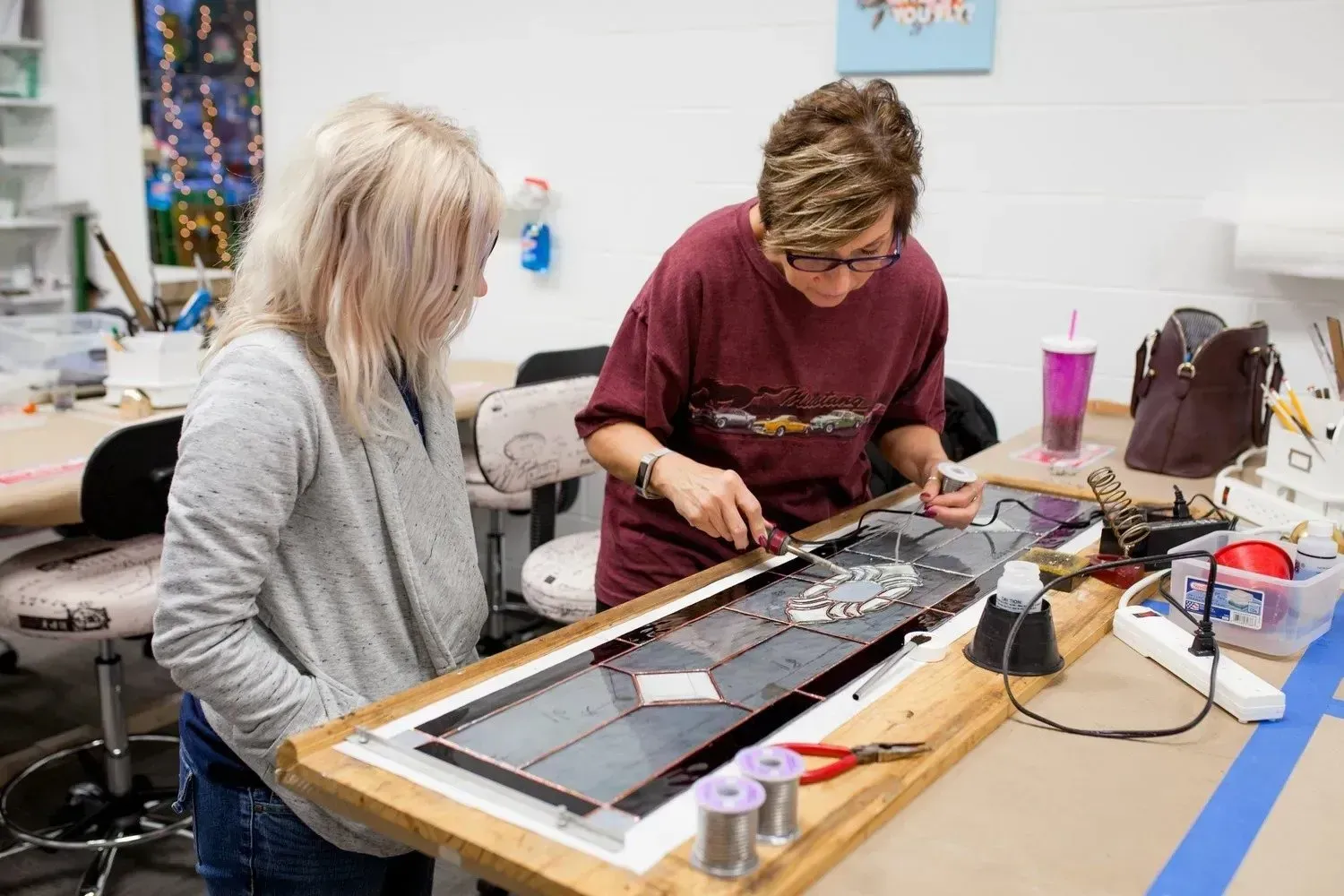 Two people working on a stained glass project in a workshop. One is soldering, the other watches.