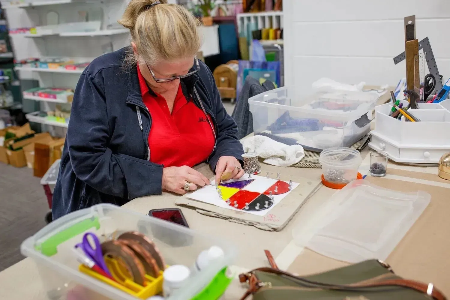 Woman in glasses working on a colorful mosaic art project at a table in a well-lit art studio.