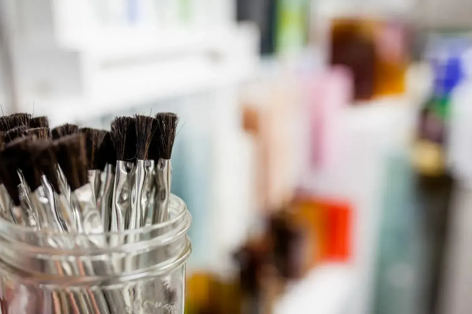 Paintbrushes in a clear glass jar, with a blurred background of colorful containers.