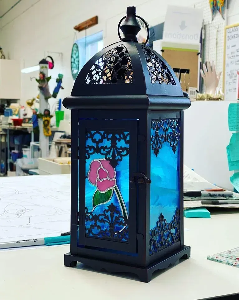 Black lantern with blue stained glass and a pink rose, set on a table.