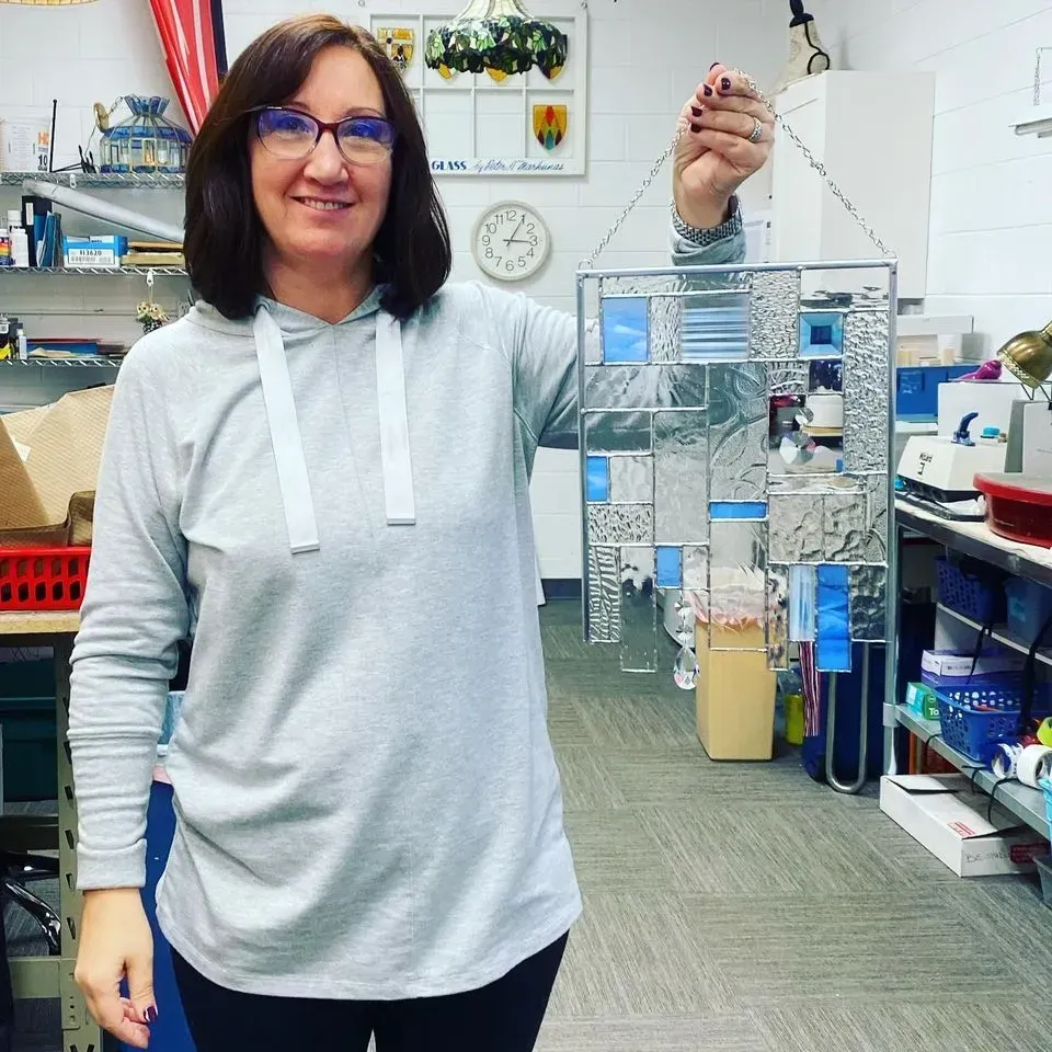 Woman in a grey hoodie holding up a piece of clear and blue stained glass art in a workshop setting.