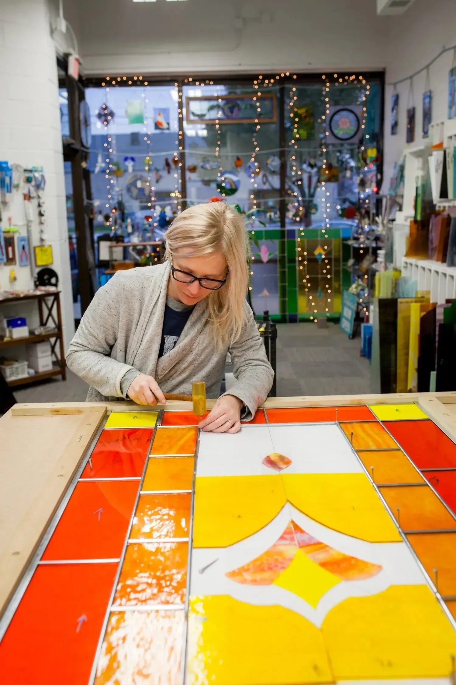 Woman working on stained glass art, using tools in a brightly lit studio.