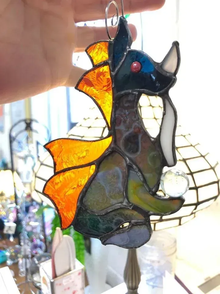 Stained glass dragon ornament with orange wings, blue eye, and multicolor body, held by a hand.