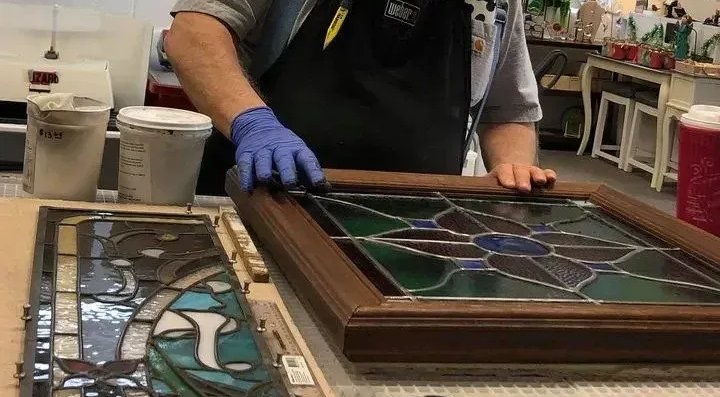 Person in gloves works on a stained glass panel, set on a workbench.