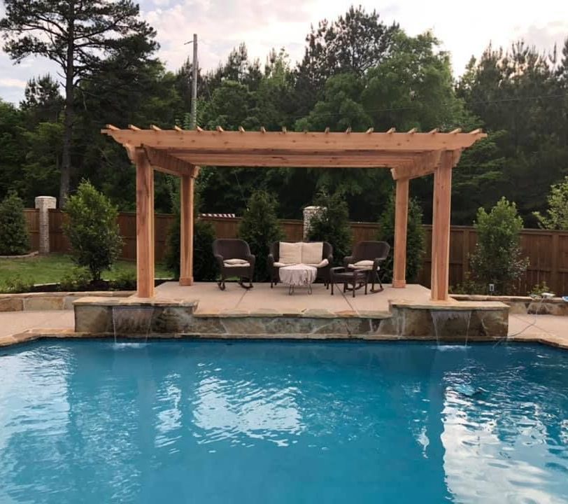 There is a pergola overlooking a swimming pool with chairs and a table.