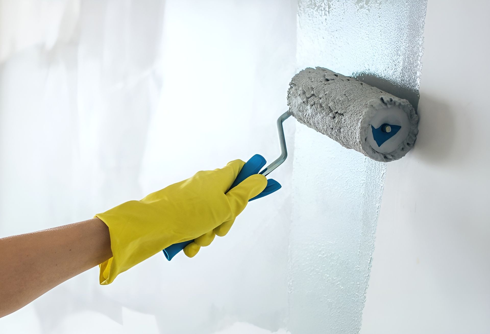 Person Wearing Yellow Gloves Painting a White Wall with A Blue Paint Roller — Frenchy’s Plaster & Paint in Palmerston, NT