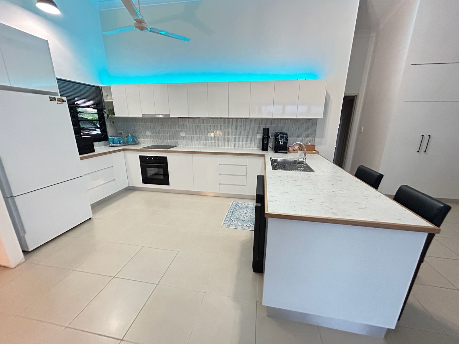 Modern white kitchen with a peninsula counter, black stools, and blue under-cabinet lighting. — Frenchy’s Plaster & Paint in Bellamack, NT