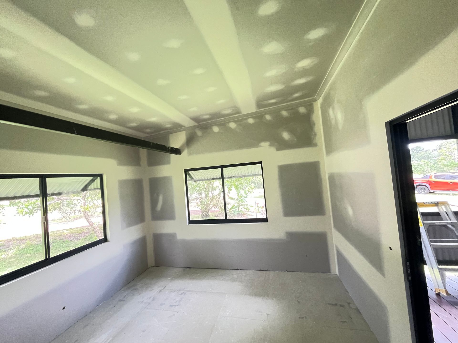 A room under construction with unfinished drywall, taped seams, and two windows looking out onto a bright outdoor area. — Frenchy’s Plaster & Paint in Bellamack, NT