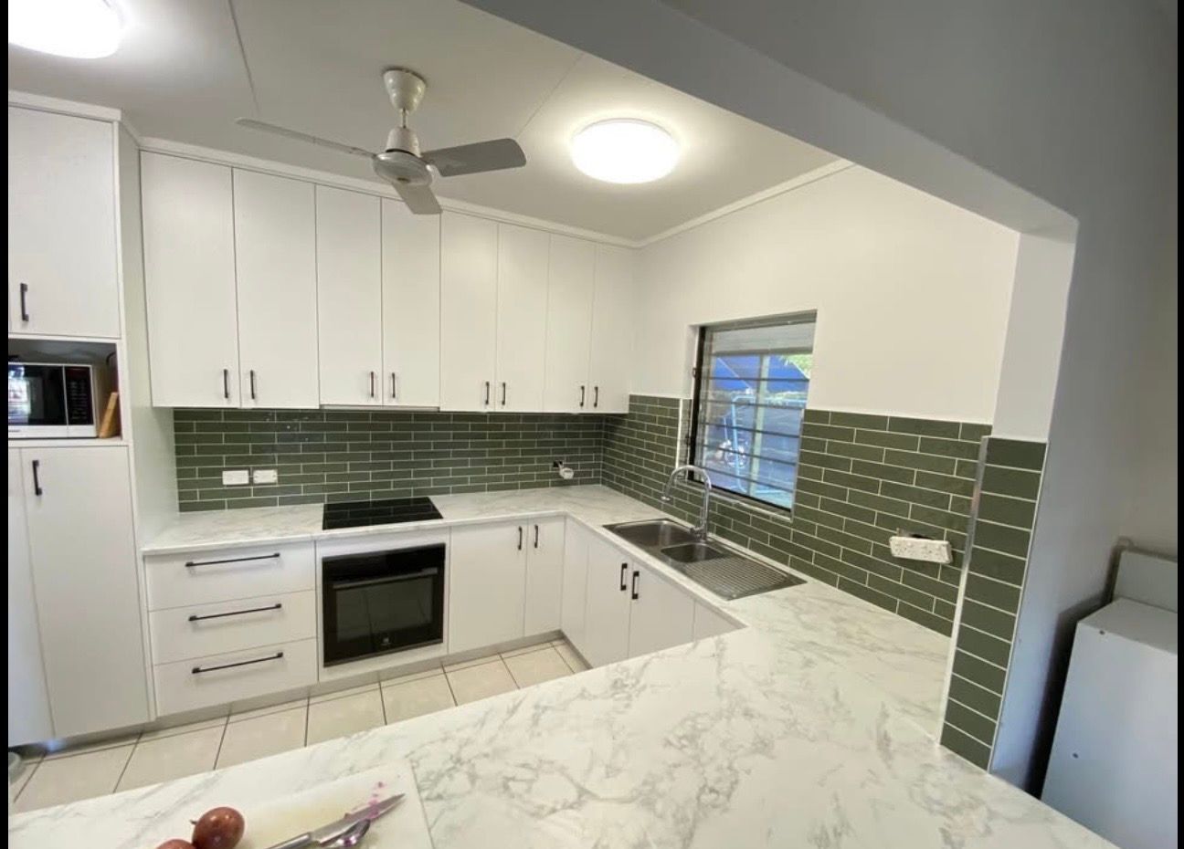 A kitchen featuring white cabinets, a marble-patterned countertop, and a green tiled backsplash with a central window. — Frenchy’s Plaster & Paint in Bellamack, NT