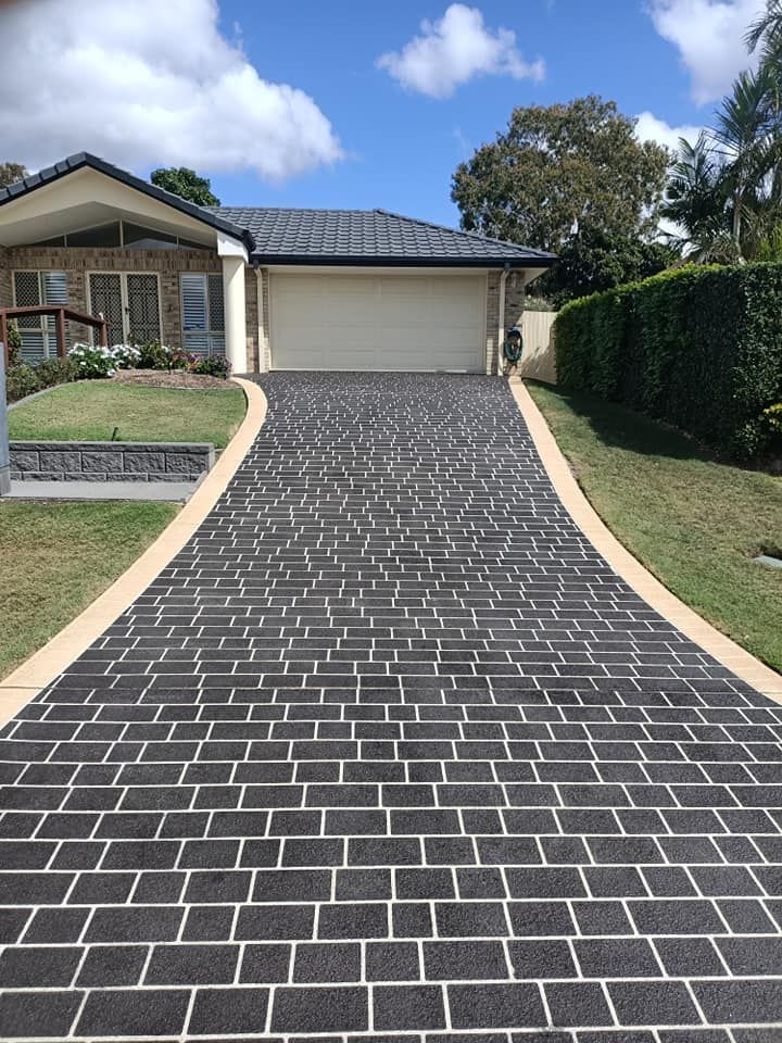 Brick-patterned driveway leading to a light-colored garage, bordered by tan concrete and green grass, with a house in the background.