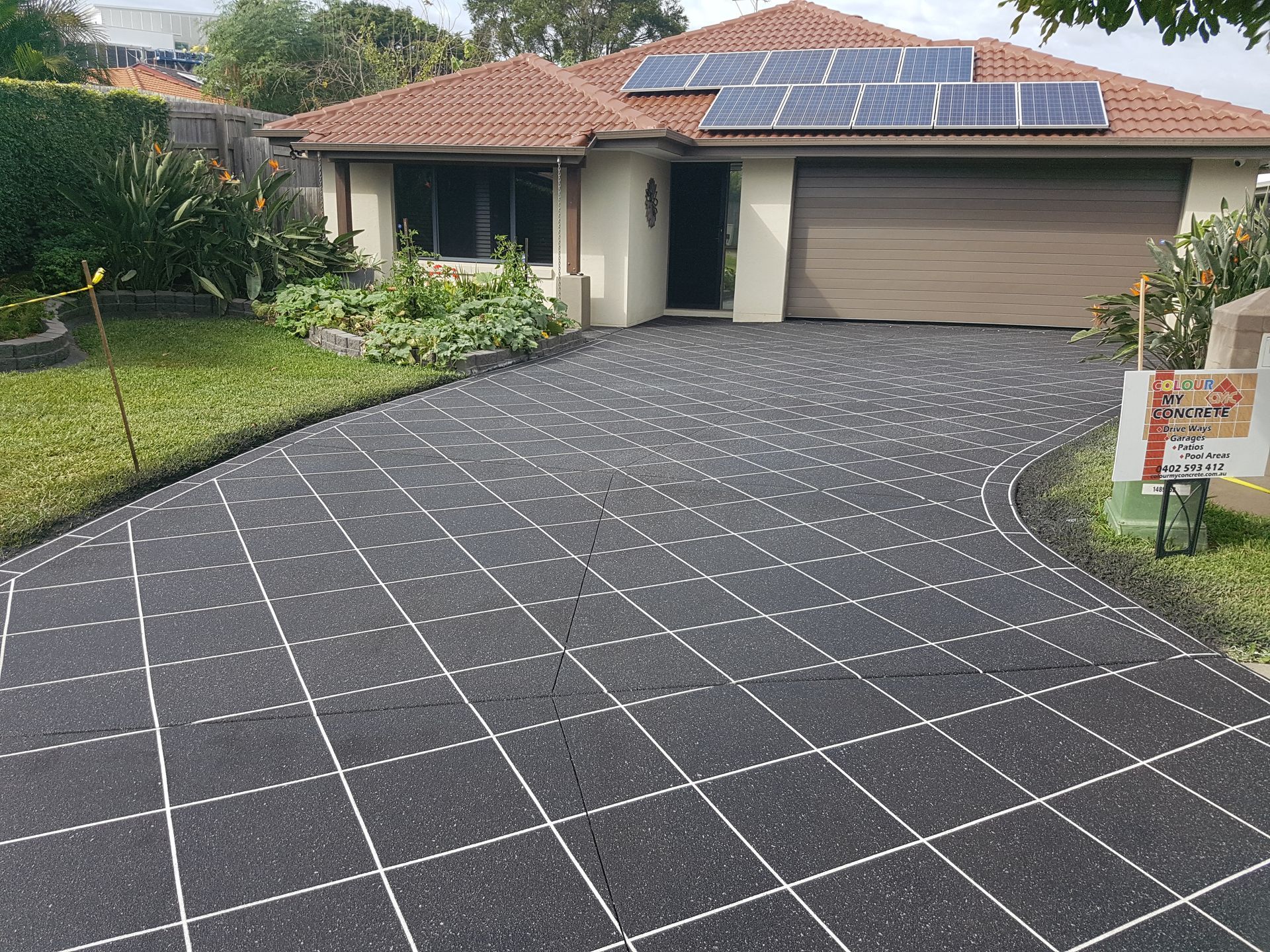 House with solar panels, a grid-patterned driveway, and a closed garage door. Green lawn and vegetation surround the house.