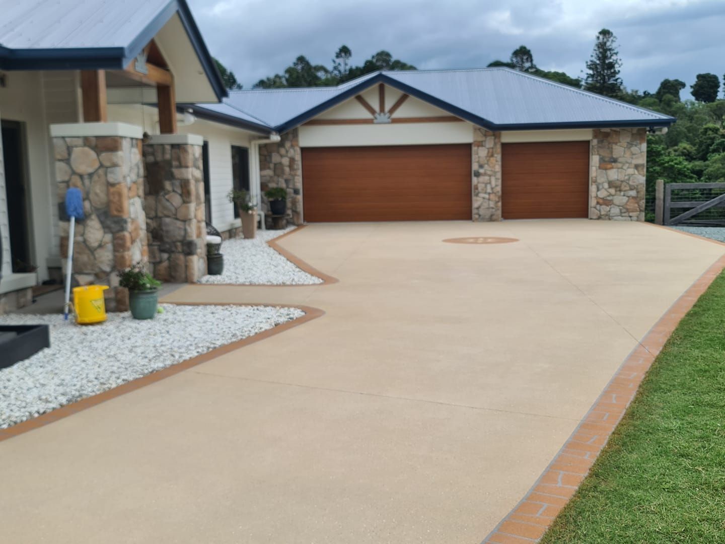 A beige concrete driveway leads to a two-car garage with brown doors, bordered by white gravel gardens and a stone pillar.