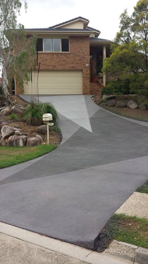 Two-story brick house with gray driveway. Mailbox in front yard. Trees and bushes surround.