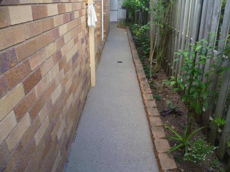Narrow concrete pathway next to a brick wall and a garden with a wooden fence.