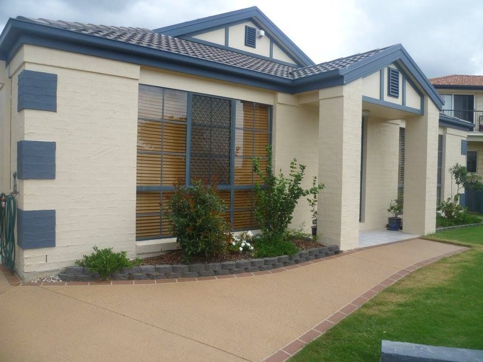 Beige house with blue trim, brown roof, and concrete pathway. Green lawn and shrubs in front.