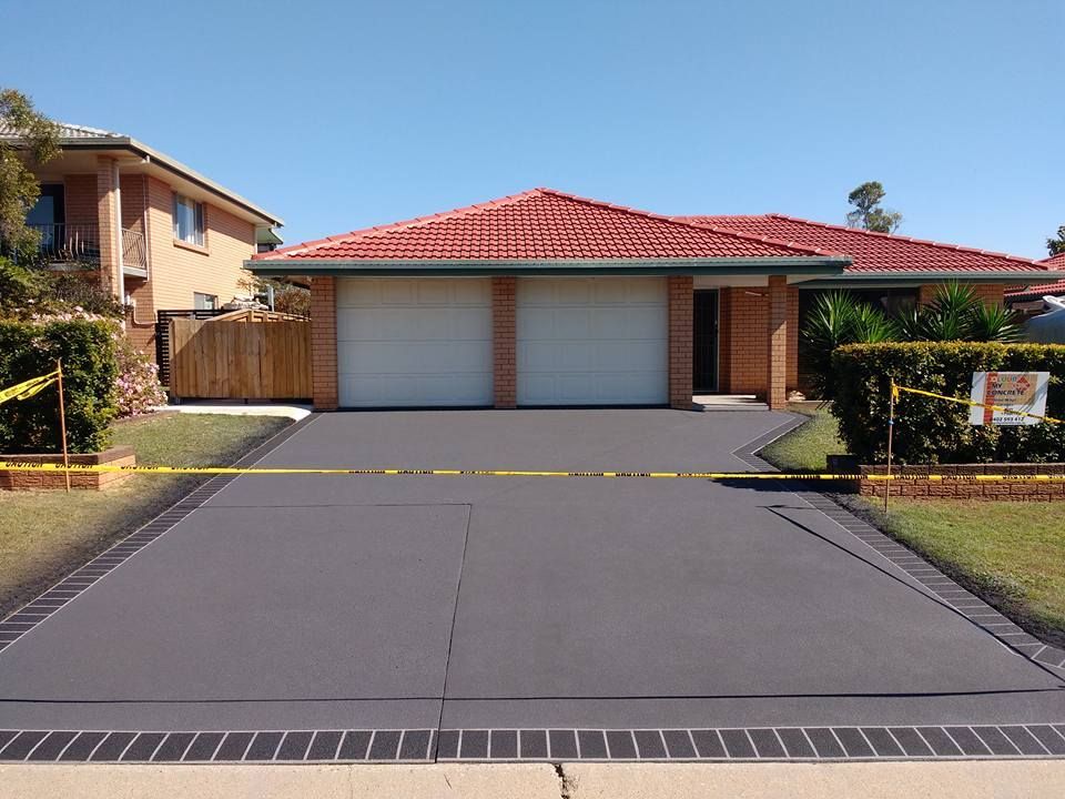 Dark gray driveway in front of a brick house with a red tile roof and two white garage doors, bordered by brick.
