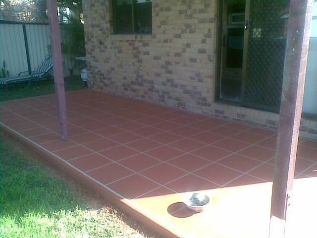 Red tiled patio next to a brick wall and green lawn, under a covered porch.