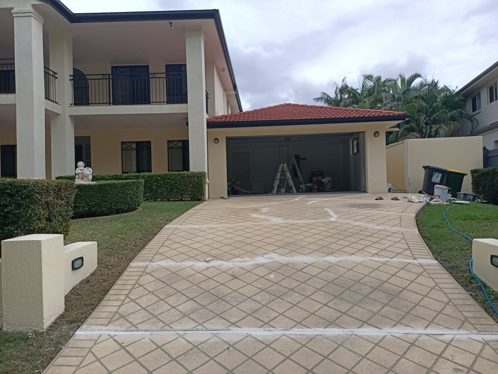 Beige house with a paved driveway leading to an open garage. Green hedge and cloudy sky visible.