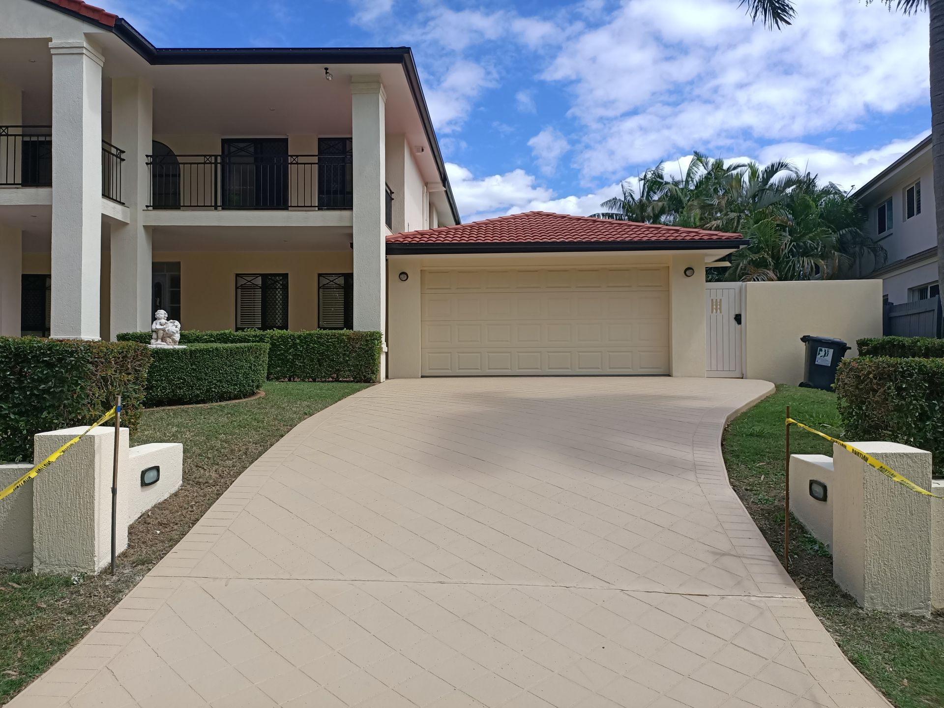 Two-story house with a driveway. Beige facade, red-tiled roof, and a garage door. Blue sky with clouds in the background.