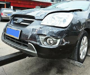 Damaged black car front, showing collision damage to the bumper, fender, and hood. Car on a tow lift.