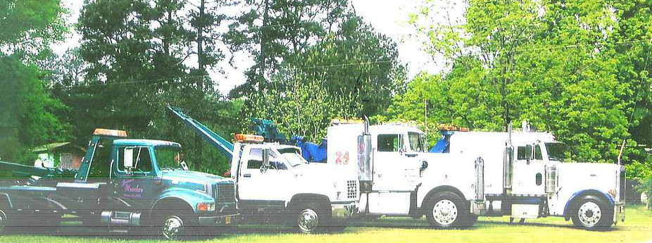 Three tow trucks parked in front of trees. One is green, two are white.