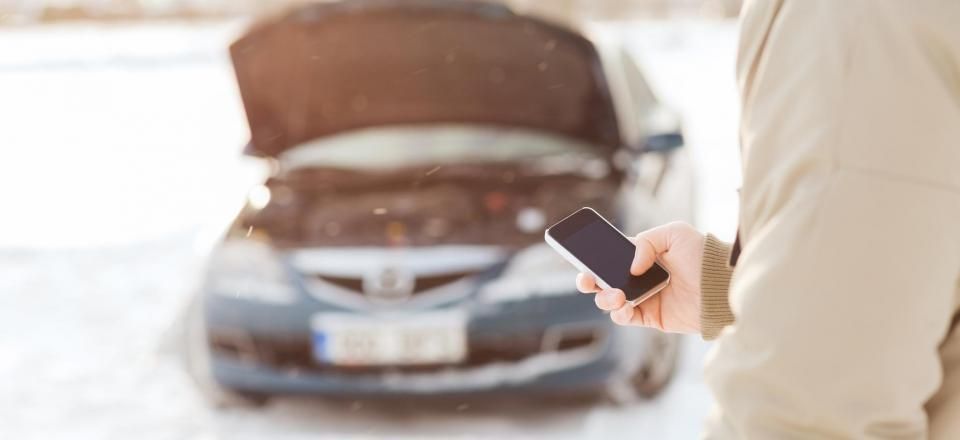 A person holding a phone near a car with an open hood, on a snow-covered road.