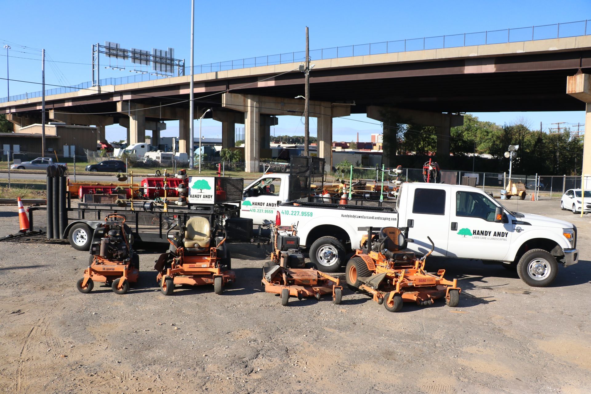 A row of lawn mowers are parked in a parking lot under a bridge.