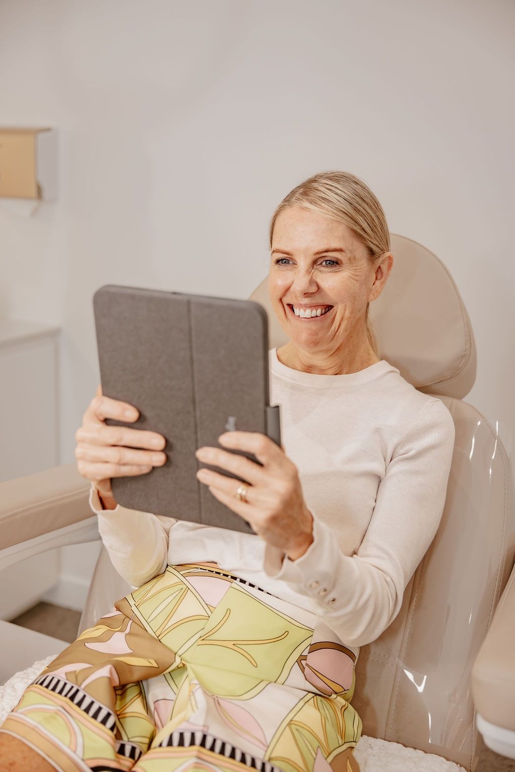 A Woman is Sitting in a Chair Holding a Tablet and Smiling — Gorjess Aesthetics in Corrimal, NSW