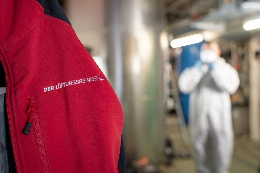 An einer Wand hängt eine rote Jacke , im Hintergrund ist ein Mann in einem weissen Anzug zu sehen.