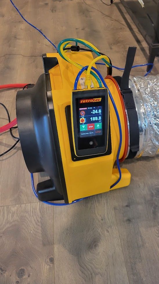 A yellow industrial air mover on a wood floor with a small digital controller screen attached and a flexible silver duct.
