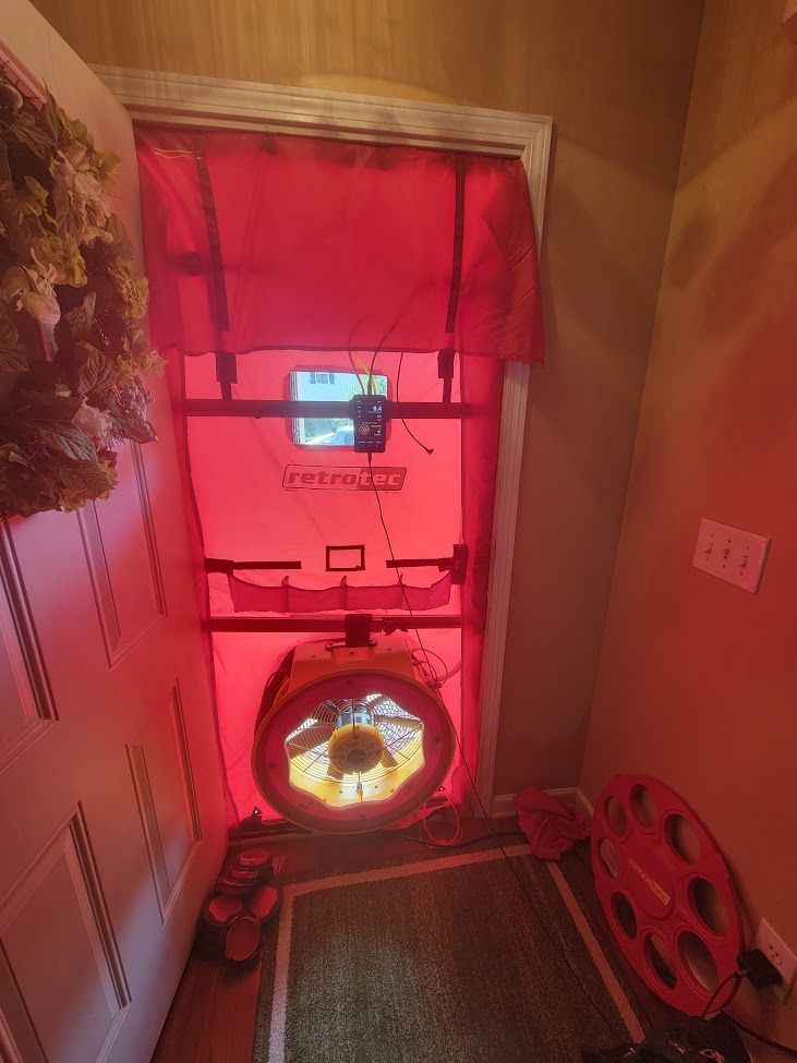 A blower door test setup with a red panel sealed in a doorway, featuring a large circular fan at the bottom.